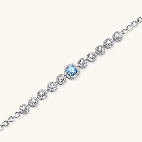 Sterling silver tennis bracelet with round halo-set moissanite stones and blue center stone on cream background.